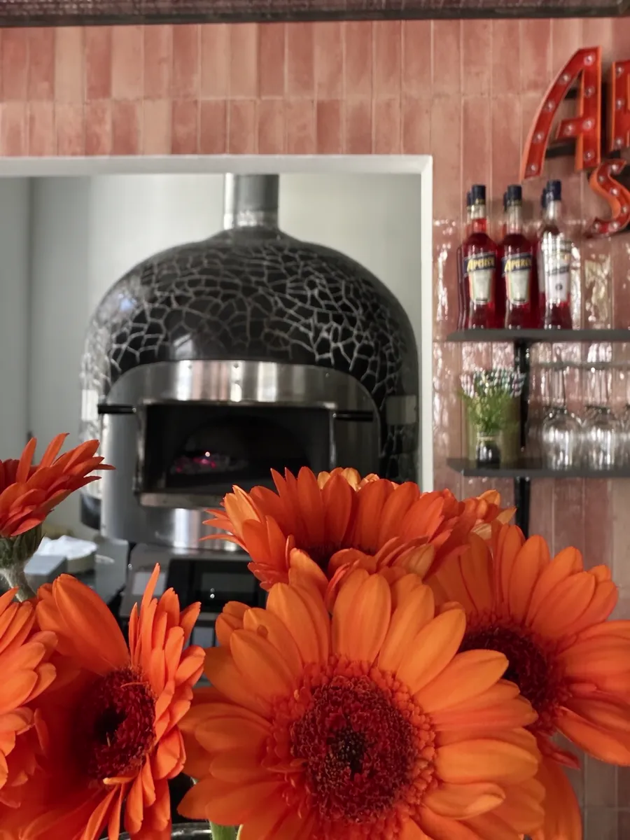 Der Pizzaofen von Fiammetta mit schwarzem Mosaik, davor orange Gerbera, rechts Aperol-Flaschen.