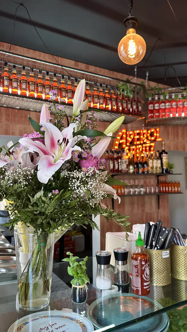 Tresen mit Lilien und Gerbera, dahinter das rote Aperol-Spritz-Licht und Flaschenregale.